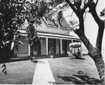 A black and white picture of a pathway and large tree near the Kirribilli House. Two people are sitting outside under an umbrella on the gardens outdoor furniture