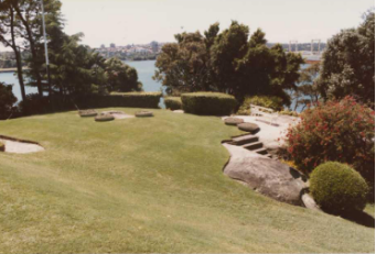 A picture of Kirribilli House gardens with areas of rock pavements, bushes, stairs and large trees and the harbour surrounding 