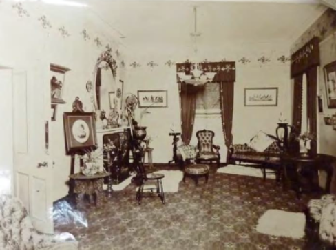 A faded sepia toned image of a room with decorated floors and walls, long draping curtains above two windows and furniture, pillows, rugs and trinkets all around 