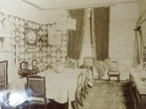 A sepia toned picture of a dining room with long draping curtains by the window, chairs by the table and around the room and art hanging from the decorated walls