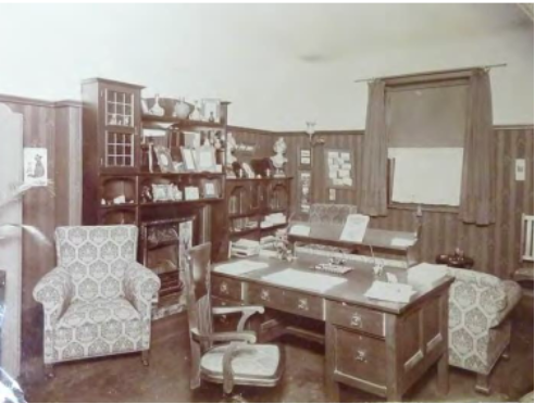 A sepia toned image of furniture within Kirribilli House such as arm chairs, study desk with papers on top, cabinet with trinkets in a room with walls partially covered in striped wallpaper