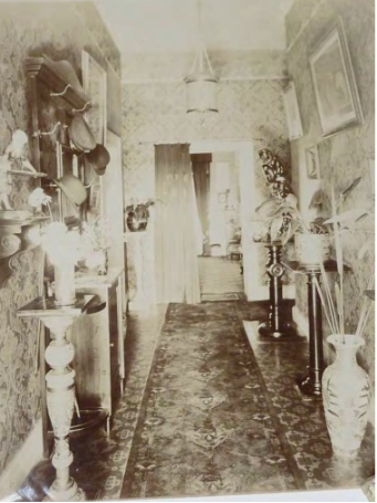 A sepia tone image of a hallway in Kirribilli House with designed floors and walls, hanging art, vases and pot plants on long thin side tables and cabinets along its sides and a curtain leading into the next room
