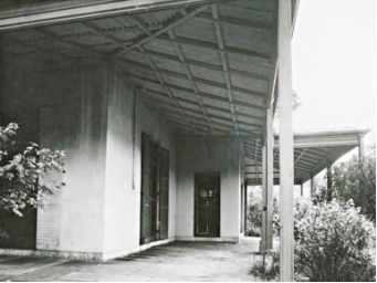 A black and white picture of the Kirribilli House front entry porch with bushes by the columns and the windows shutters closed