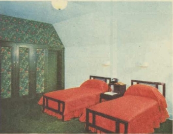 A picture of a bedroom with two red single beds on simple wood bed frames with a bed side table in between both beds and a decorated wardrobe towards the left