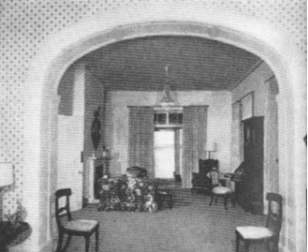 A black and white picture of the Kirribilli House drawing room with minimal furniture, a fireplace and an arch entry way